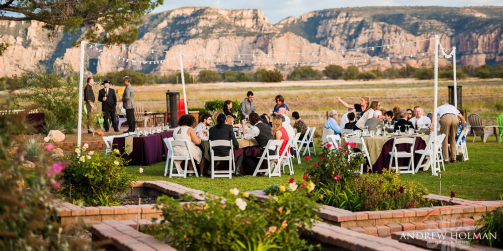 An outdoor event at Mesa Grill Sedona, showcasing guests seated at white linen-covered tables arranged on a lush lawn area. The venue features a fire pit, elegant brickwork, and well-tended garden beds with blooming flowers. String lights are stretched above the diners, adding a festive ambiance. In the background, majestic red rock formations rise against a clear blue sky, offering a stunning natural backdrop to the gathering. The image captures a lively atmosphere with guests engaged in conversation, dining, and enjoying the scenic views.