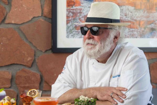 Chef Mercer Mohr seated at a table adorned with a variety of plated dishes, wearing sunglasses and a white hat, with a framed photo of Sedona’s red rocks in the background.