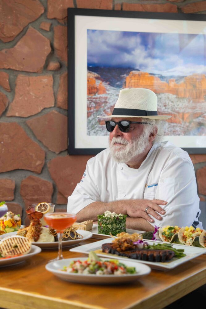 Chef Mercer Mohr seated at a table adorned with a variety of plated dishes, wearing sunglasses and a white hat, with a framed photo of Sedona’s red rocks in the background.