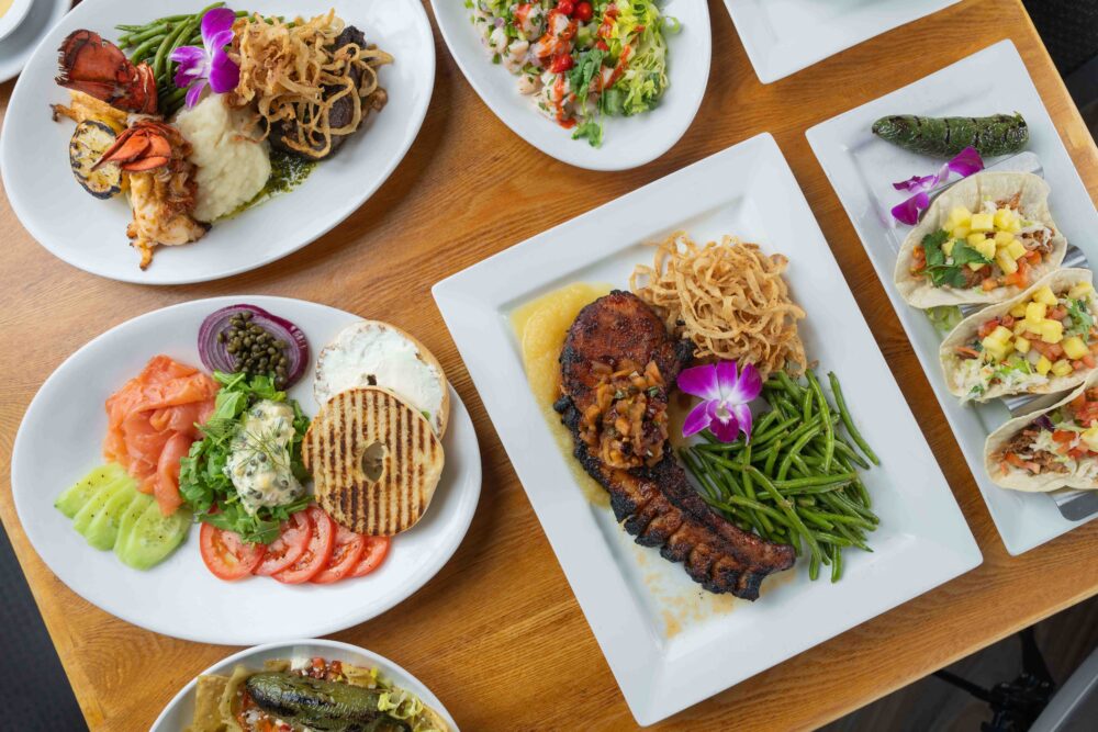 A variety of beautifully plated dishes displayed on a wooden table, including smoked salmon with bagel, tacos, tomahawk pork chop with green beans, steak and lobster, and a fresh salad.